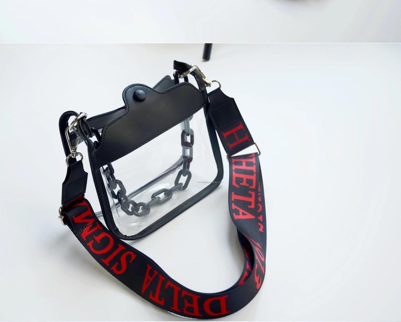 May include: A clear, rectangular crossbody bag with a black top and chain detail. The black strap features red text that reads "DELTA SIGMA THETA". The bag has a black chain inside.