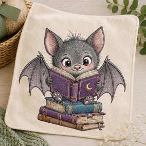 May include: A cream-colored fabric square featuring an embroidered illustration of a gray bat with large eyes and pink cheeks. The bat is sitting on a stack of two books while holding an open purple book with a gold crescent moon on the cover.