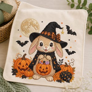 May include: A square off-white fabric piece featuring an embroidered design of a light brown rabbit wearing a black witch hat with an orange plaid bow and a black cape. The rabbit holds a jack-o'-lantern bucket filled with colorful candies.