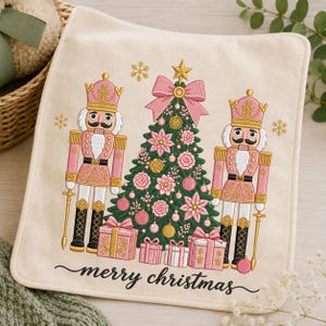 May include: A cream-colored fabric square featuring an embroidered design of a green Christmas tree decorated with pink ornaments and a large pink bow, flanked by two pink nutcrackers. Below the tree are pink gift boxes and the text merry christmas.