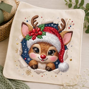 Op de afbeelding: Een vierkant, crèmekleurig stuk stof met een borduursel van een rendier dat een rood-witte kerstmuts draagt. Het rendier wordt omlijst door een donkerblauwe, cirkelvormige uitsnede met witte sneeuwvlokken, groene hulstbladeren en rode bessen.