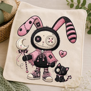 May include: A square off-white fabric patch featuring an embroidered design of a black and pink rabbit character with button eyes, wearing a cape and sneakers, holding a crescent moon wand, accompanied by a small black cat with a skull collar.