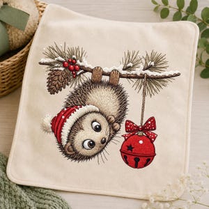 May include: A square cream-colored fabric cloth featuring an embroidered illustration of a hedgehog wearing a red and white winter hat, hanging upside down from a pine branch with a pinecone, red berries, and a large red jingle bell with a bow.