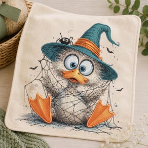 May include: A square off-white fabric piece featuring an embroidered illustration of a fluffy duckling wearing a teal witch hat with an orange band. The duckling has large blue eyes, an orange beak, and orange feet, entangled in a black spider web.