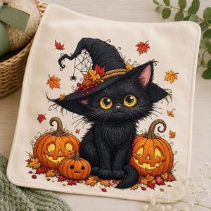 May include: A square off-white fabric item featuring a black kitten wearing a pointed witch hat adorned with autumn leaves and berries. The kitten sits between two large jack-o'-lanterns and one small pumpkin, surrounded by scattered orange and red maple leaves.