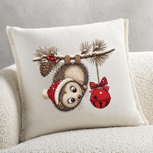 May include: A square off-white throw pillow with fringe edges featuring a printed illustration of a hedgehog wearing a red and white winter hat, hanging upside down from a pine branch with red berries, a pinecone, and a red jingle bell ornament.