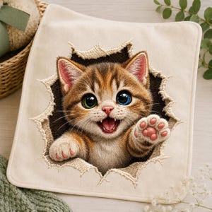 Op de afbeelding: Een vierkante, crèmekleurige stoffen patch met een geborduurd ontwerp van een bruine cyperse kitten die door het oppervlak lijkt te breken. De kitten heeft één groen oog en één blauw oog, een open mond en één opgeheven pootje.