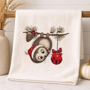 May include: A white kitchen towel featuring a printed illustration of a brown hedgehog wearing a red and white winter hat, hanging from a snowy pine branch with red berries and a pinecone, while holding a large red jingle bell with a bow.