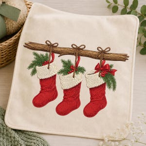 Peut inclure: Un article en tissu carré de couleur blanc cassé présentant une illustration imprimée de trois chaussettes de Noël rouges avec des revers en fausse fourrure blanche suspendues à une branche en bois marron, décorées de pin et de baies.