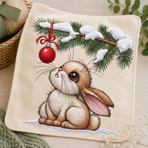 May include: A square cream-colored fabric piece featuring an embroidered design of a brown rabbit sitting in snow, looking up at a red Christmas ornament hanging from a snow-covered pine branch with green needles and white snow accents.