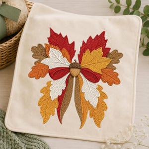 May include: A square cream-colored fabric napkin featuring an intricate machine embroidery design of autumn leaves in shades of red, orange, yellow, and brown, centered with a small brown acorn and a red ribbon bow detail.