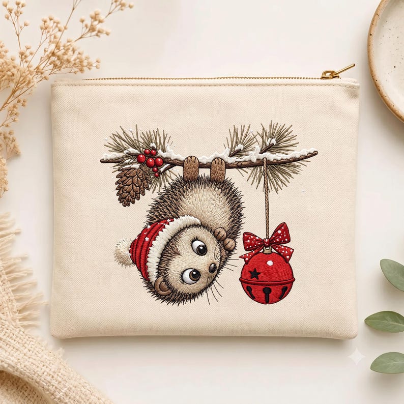 May include: A cream-colored canvas zipper pouch featuring an embroidered illustration of a hedgehog wearing a red and white winter hat, hanging upside down from a pine branch with red berries, a pinecone, and a red jingle bell with a bow.