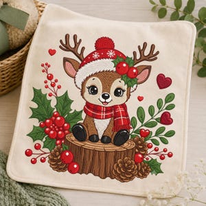 May include: A square off-white fabric piece featuring an embroidered design of a brown fawn sitting on a tree stump. The fawn wears a red winter hat with a pom-pom, a red plaid scarf, and is surrounded by holly leaves, red berries, and pinecones.
