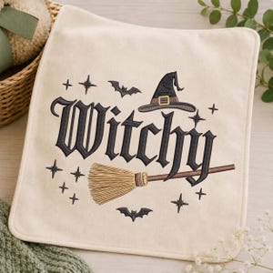 May include: A square cream-colored fabric item featuring black embroidered text that reads Witchy. The design includes a black witch hat with a gold buckle, a brown broomstick, two black bats, and several small black star-shaped accents.