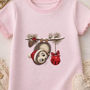 May include: A light pink short-sleeved t-shirt featuring a graphic of a hedgehog wearing a red and white winter hat, hanging upside down from a pine branch with a red jingle bell, pinecones, and red berries.