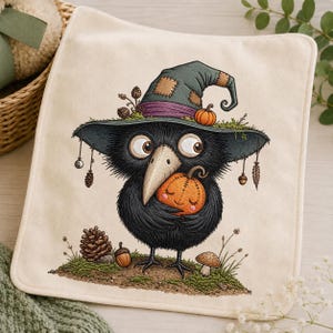 Può includere: Un panno in tessuto quadrato color bianco sporco con un'illustrazione dettagliata di un uccello nero che indossa un grande cappello da strega verde con una fascia viola. L'uccello tiene in mano una piccola zucca arancione, circondato da pigne, ghiande e un fungo.