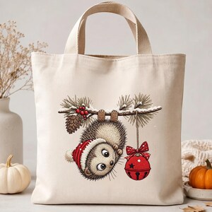 May include: A cream-colored canvas tote bag featuring a graphic illustration of a hedgehog wearing a red winter hat, hanging upside down from a pine branch with red berries, a pinecone, and a red jingle bell ornament with a polka dot bow.