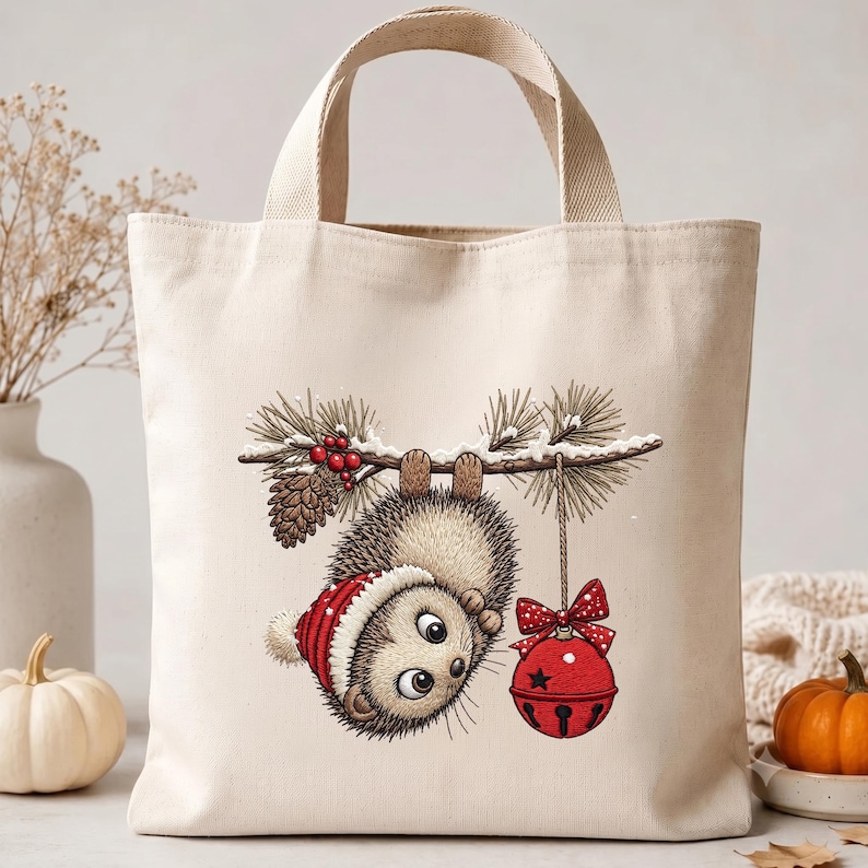 May include: A cream-colored canvas tote bag featuring a graphic illustration of a hedgehog wearing a red winter hat, hanging upside down from a pine branch with red berries, a pinecone, and a red jingle bell ornament with a polka dot bow.
