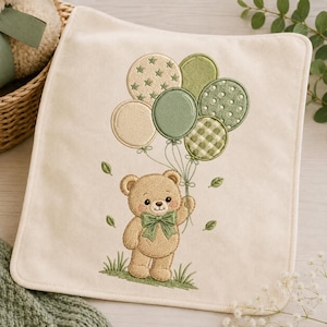 Puede incluir: Un cuadrado de tela color crema con un bordado de un oso de peluche marrón claro que lleva una pajarita verde. El oso sostiene un racimo de seis globos en varios tonos de verde y crema, decorados con patrones de estrellas, puntos y cuadros.