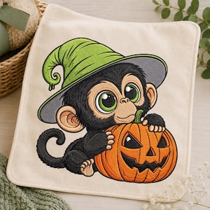 Puede incluir: Un paño de tela cuadrado de color blanco roto con un diseño bordado de un mono negro de ojos verdes grandes que lleva un sombrero de bruja verde brillante. El mono sostiene una calabaza naranja con una cara tallada y un tallo verde.