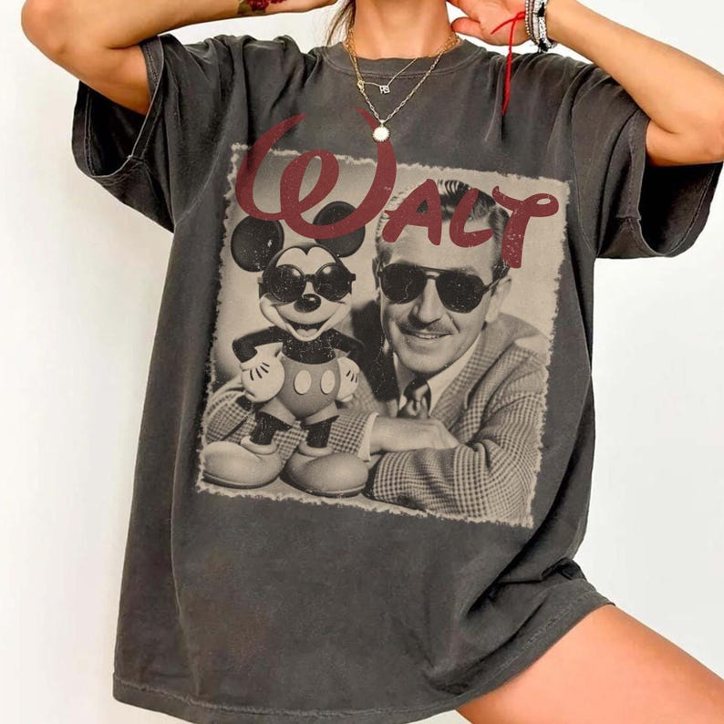 May include: A charcoal gray short-sleeve t-shirt featuring a graphic print of a man and a cartoon mouse both wearing sunglasses. The text "WALT" is printed in a dark red script font above the figures, which are depicted in a sepia-toned square.