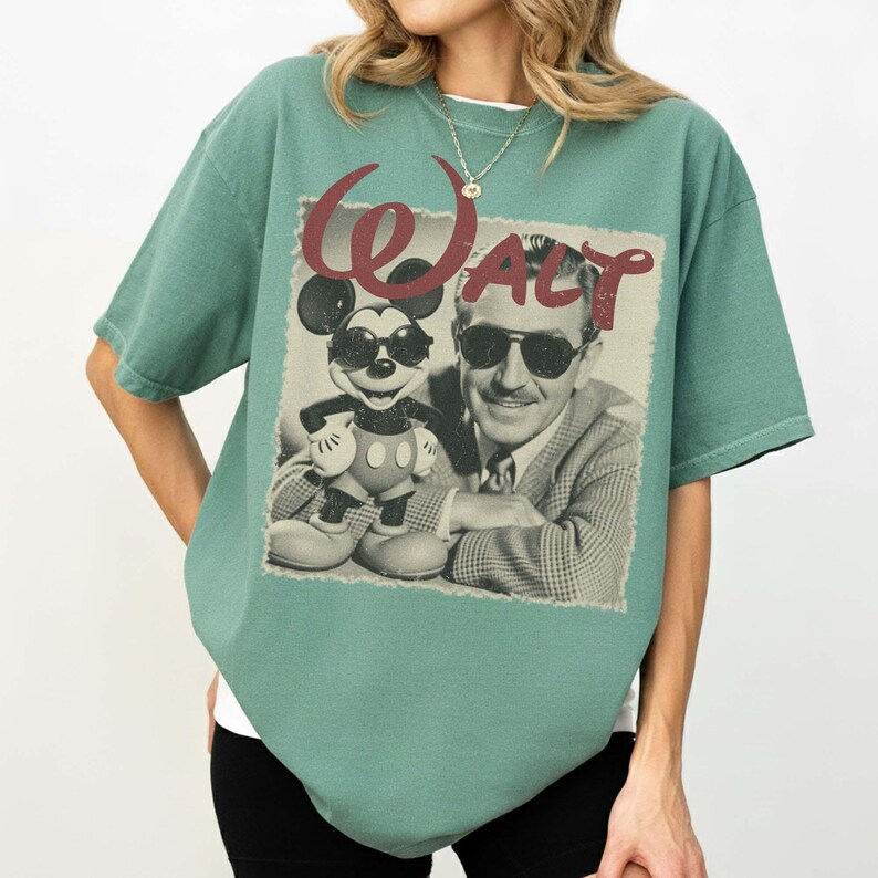 May include: A sage green short-sleeve t-shirt featuring a graphic print of Walt Disney and Mickey Mouse both wearing sunglasses. The word Walt is printed in a large, distressed red font above the black and white image of the two figures.