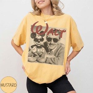 May include: A mustard yellow short-sleeve t-shirt featuring a graphic print of a man in a suit and a cartoon mouse, both wearing sunglasses. The text "Walt" is printed in a large, distressed red font above the figures on the front of the shirt.