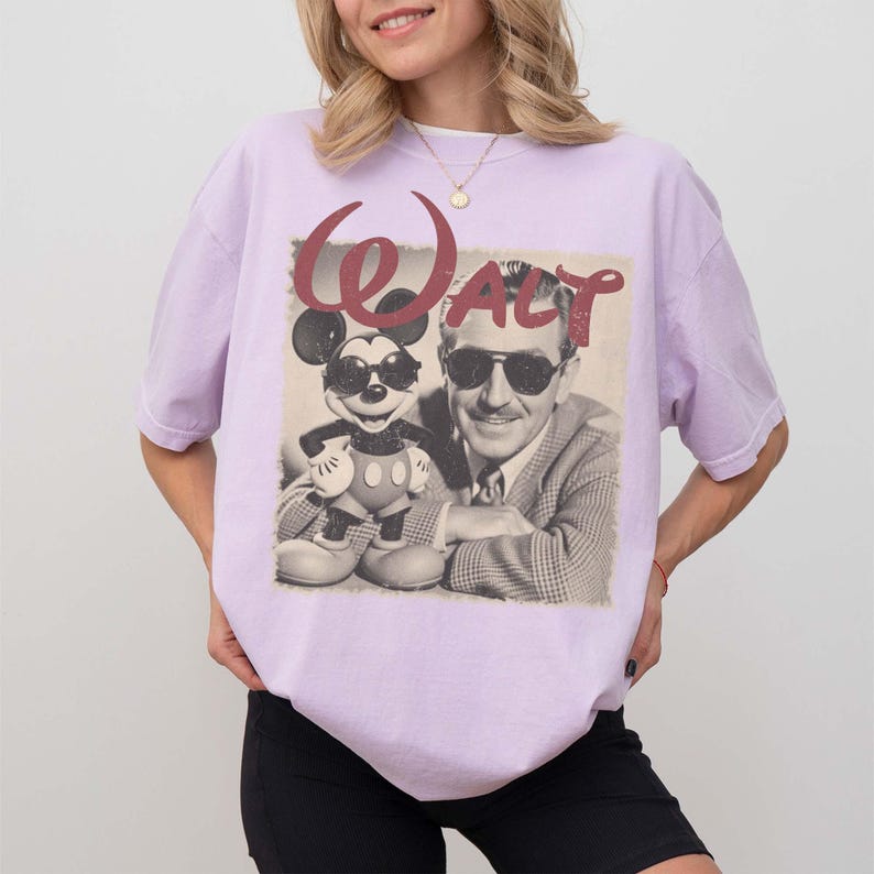 May include: A light purple short-sleeve t-shirt featuring a graphic print of Mickey Mouse and Walt Disney both wearing sunglasses. The word Walt is printed in a large, distressed red font above the black and white image of the two figures.