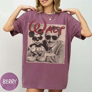 May include: A berry-colored short-sleeve t-shirt featuring a graphic print of Mickey Mouse and Walt Disney both wearing sunglasses. The text "WALT" is printed in a large, stylized red font above the figures. The shirt has a crew neckline.