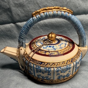 May include: An ornate teapot with a cream-colored base, blue handle, and gold accents. The lid features a gold knob and a decorative blue and white pattern. A gold chain secures the lid to the spout.