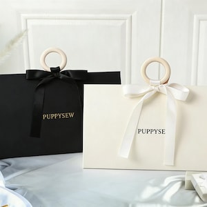 Op de afbeelding: Twee cadeauzakken, één zwart en één crème, met houten ringgrepen en satijnen strikken. Op de tassen staat het woord "PUPPYSEW". Een klein geschenkdoosje is zichtbaar op de achtergrond.