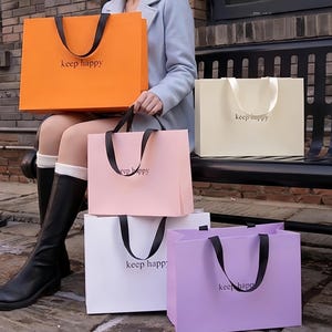 Puede incluir: Una variedad de bolsas de compras coloridas en naranja, rosa, blanco y lila. Cada bolsa tiene un asa negra y la frase "keep happy" impresa. Las bolsas están apiladas y sostenidas por una persona.