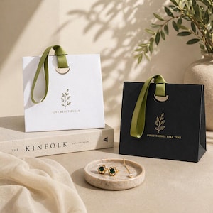 Puede incluir: Dos bolsas de regalo, una blanca y otra negra, con asas de cinta verde oliva. La bolsa blanca tiene el texto "LIVE BEAUTIFULLY" y la bolsa negra tiene el texto "GOOD THINGS TAKE TIME". Ambas bolsas presentan un diseño de hoja dorada.
