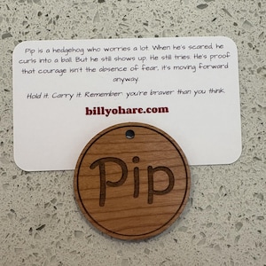 May include: A wooden round tag with the name "Pip" engraved in a cursive font. The tag is brown and has a small hole at the top. A white card with text about courage is behind the tag.
