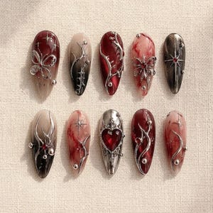 Può includere: Un set di dieci unghie finte a mandorla con una palette di colori rosso scuro, nero e crema. I design includono dettagli a farfalla in metallo 3D, motivi a viticci spinosi, stelle e una gemma centrale rossa a forma di cuore.