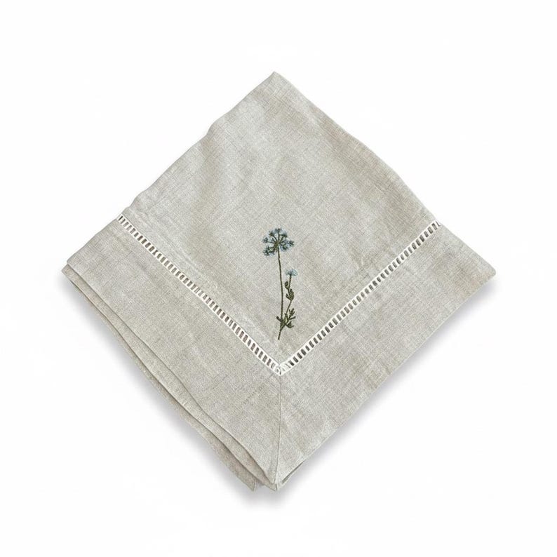 May include: A folded, pale grey linen napkin with an embroidered floral design. The napkin features a small, light blue flower with green leaves and a white, cut-out border. The napkin is square and has a soft, natural texture.