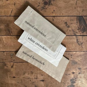 May include: Three linen napkins in neutral tones, arranged on a wooden surface. The napkins are in shades of beige and white, with embroidered details and text that reads "natural entredeux", "white entredeux", and "natural hemstitch".