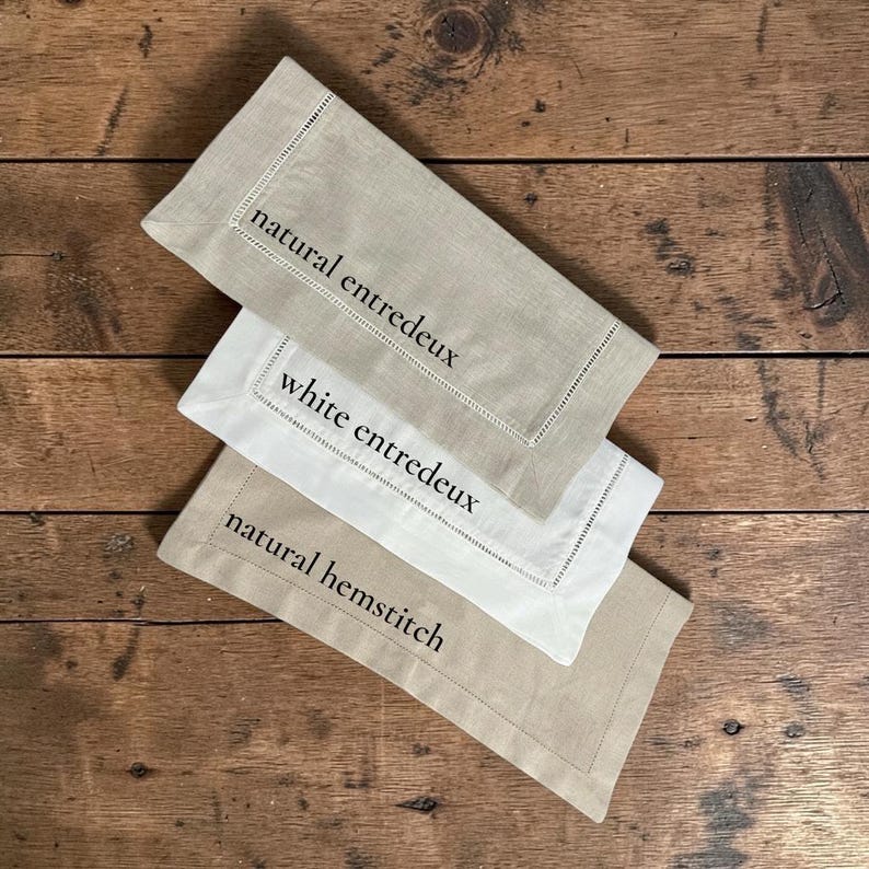 May include: Three linen napkins in neutral tones, arranged on a wooden surface. The napkins are in shades of beige and white, with embroidered details and text that reads "natural entredeux", "white entredeux", and "natural hemstitch".