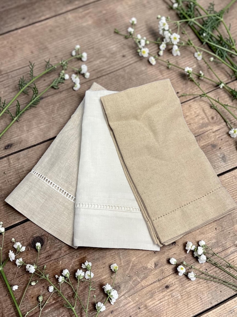 May include: Three folded linen napkins in shades of beige and cream, arranged on a wooden surface. The napkins have decorative stitched edges. White flowers and greenery are scattered around the napkins.