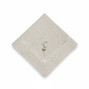 May include: A square, light beige linen napkin with an embroidered floral design. The napkin has a white, cut-out border and a small, embroidered sprig of white flowers and green leaves in the centre. The napkin is made of linen.