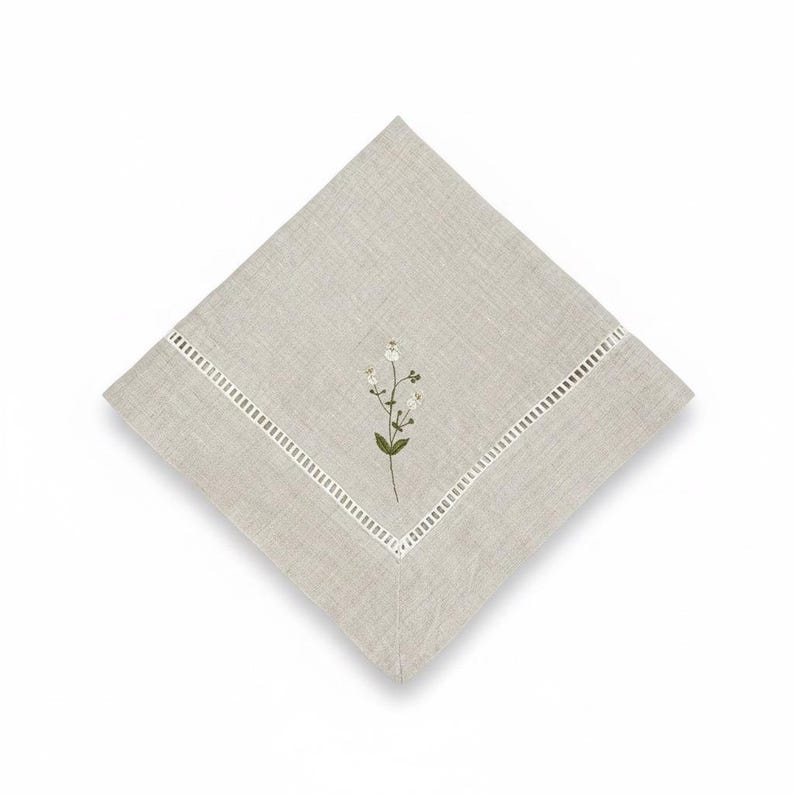 May include: A square, light beige linen napkin with an embroidered floral design. The napkin has a white, cut-out border and a small, embroidered sprig of white flowers and green leaves in the centre. The napkin is made of linen.