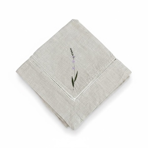 May include: A folded, pale grey linen napkin with a white, decorative border. The napkin features an embroidered lavender sprig with purple flowers and a green stem. The napkin is square and folded diagonally.