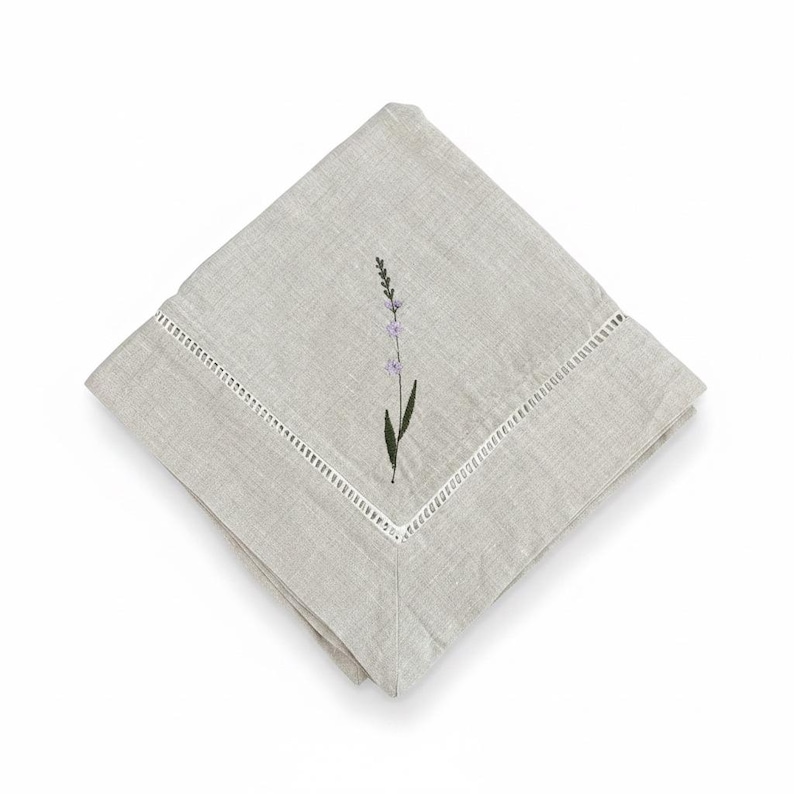 May include: A folded, pale grey linen napkin with a white, decorative border. The napkin features an embroidered lavender sprig with purple flowers and a green stem. The napkin is square and folded diagonally.