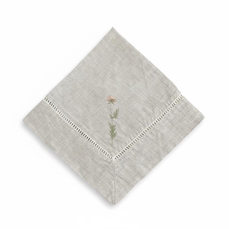 May include: A square, pale grey linen napkin featuring an embroidered flower design. The flower is a soft pink with green leaves and stem. The napkin has a white, cut-out border along one edge.