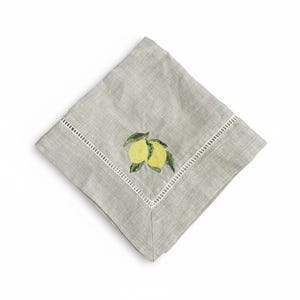 May include: A folded gray linen napkin with a white embroidered border. The napkin features an embroidered design of two yellow lemons with green leaves. The napkin is square and is set against a white background.