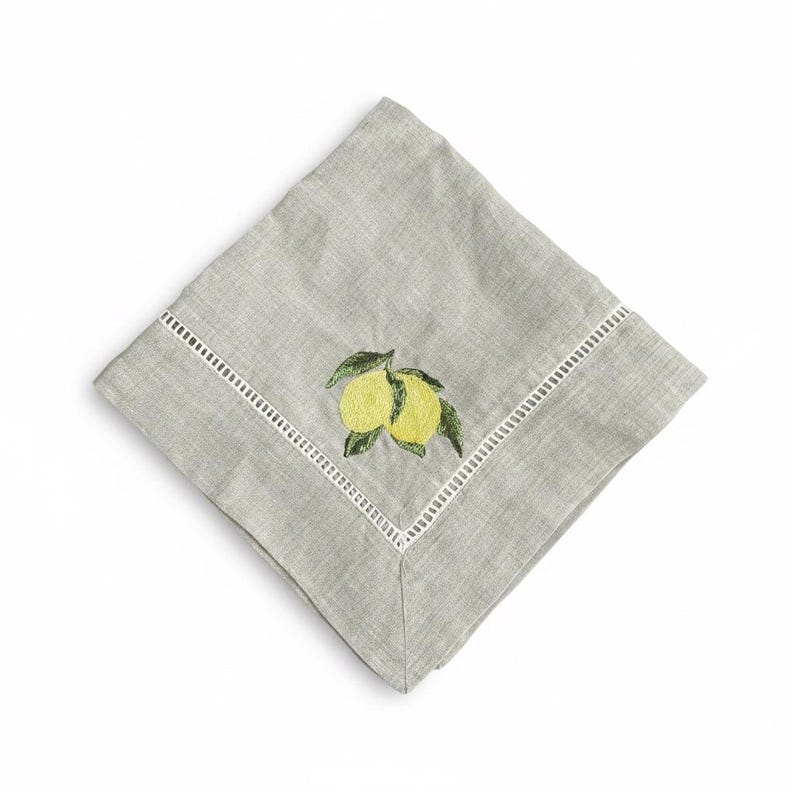 May include: A folded gray linen napkin with a white embroidered border. The napkin features an embroidered design of two yellow lemons with green leaves. The napkin is square and is set against a white background.