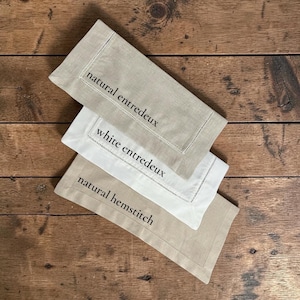 May include: Three linen napkins in neutral tones, arranged on a wooden surface. The napkins are in shades of beige and white, with embroidered details and text that reads "natural entredeux", "white entredeux", and "natural hemstitch".