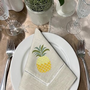 May include: A folded beige linen napkin with a yellow and green embroidered pineapple design, placed on a white plate. The napkin has a white embroidered border. The table setting includes silverware, a wine glass, and decorative items.