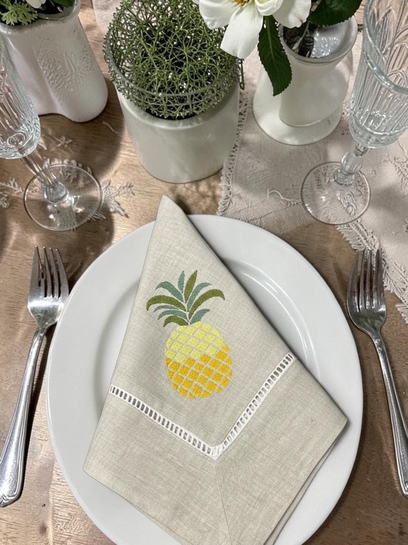 May include: A folded beige linen napkin with a yellow and green embroidered pineapple design, placed on a white plate. The napkin has a white embroidered border. The table setting includes silverware, a wine glass, and decorative items.