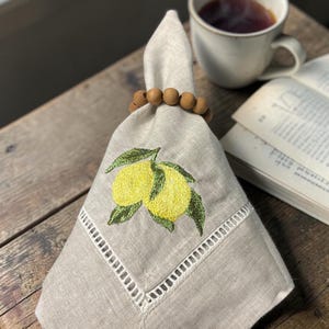 May include: A folded beige linen napkin with an embroidered lemon design, held by a wooden bead napkin ring. A cup of tea and an open book are in the background. The napkin features a decorative white border.